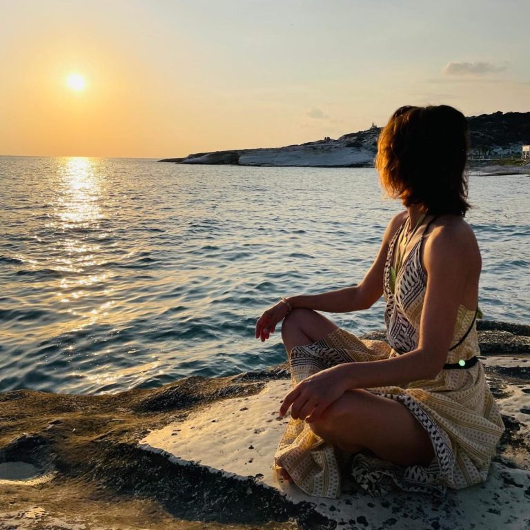 Britta Martin Körper- & Bewegungstherapeuthin Britta Martin sitzt am Meeresufer auf einem Stein und beobachtet den sanften Sonnenuntergang über dem Wasser.