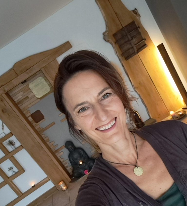 Britta Martin lächelt vor einer warmen, holzgetäfelten Wand mit Buddha-Statuette im Hintergrund.